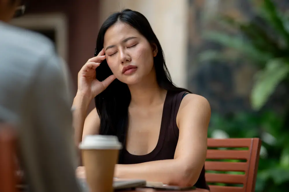 A woman touching her head with eyes closed at table.