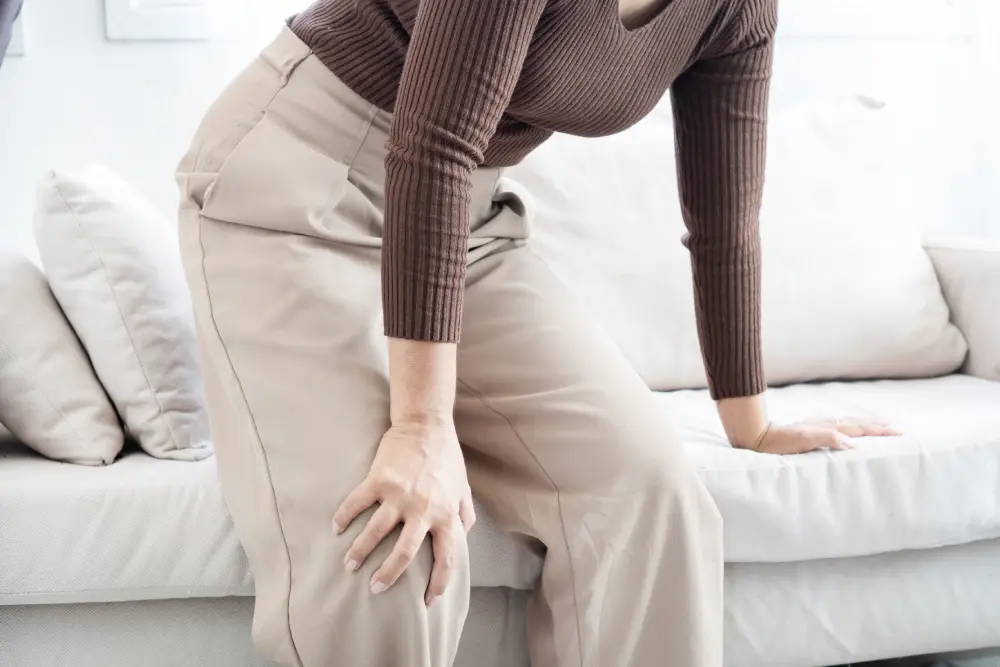 A woman holding her painful knee near a couch.